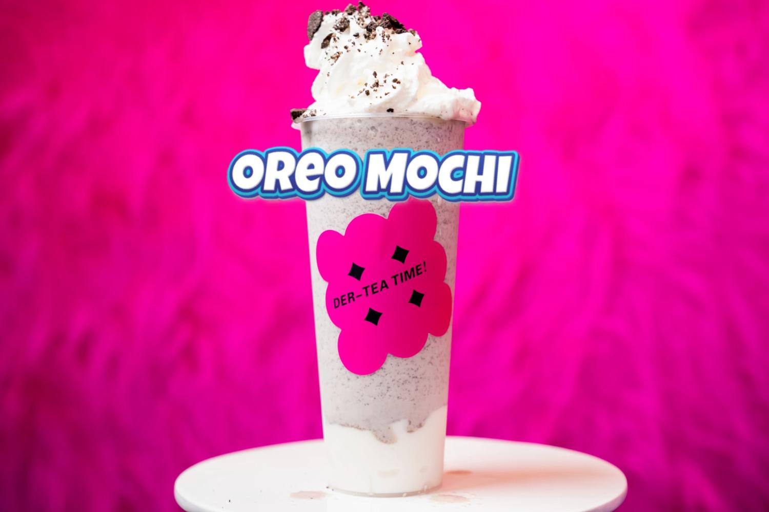 Oreo Mochi drink with whipped cream and cookie crumbs, sitting on a white table against a bright pink background.